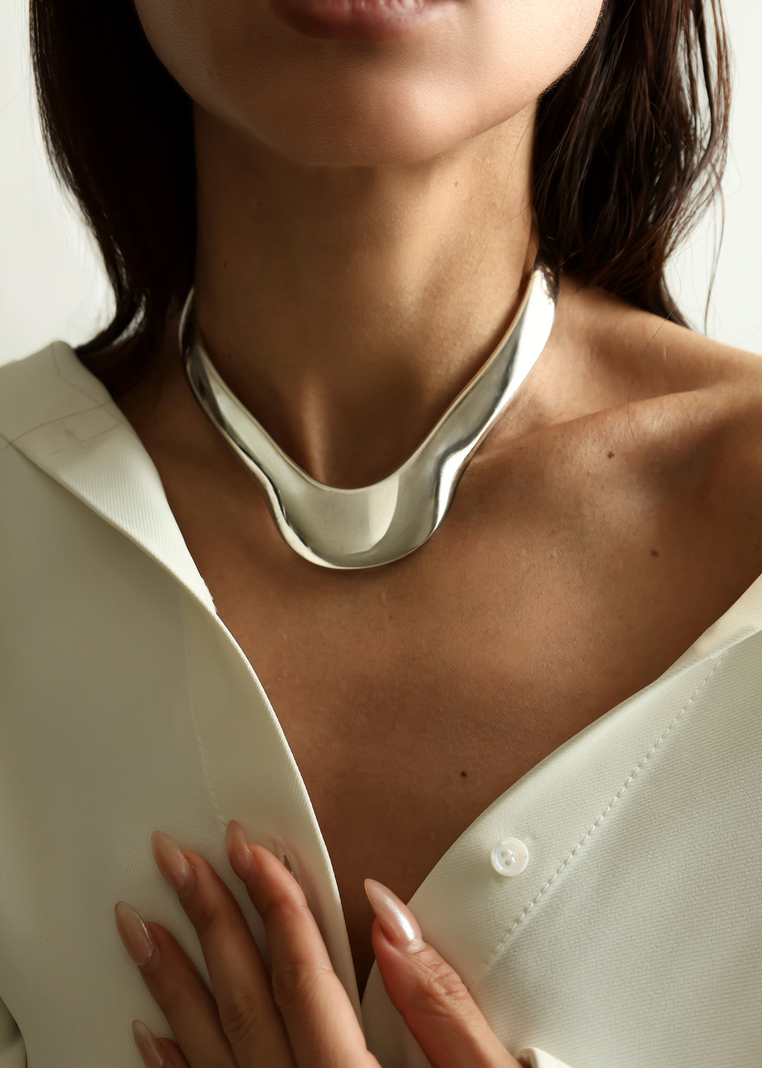 A close-up of a model wearing a bold, sculptural silver metallic choker necklace with a wide, curved collar, paired with a white collared shirt.