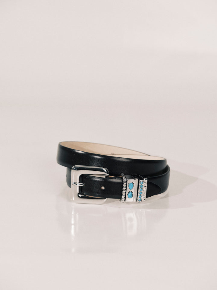 A black leather belt with a silver-tone buckle and a decorative silver-tone keeper featuring light blue stones, coiled on a white reflective surface.
