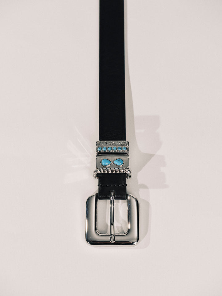 A black leather belt with a silver-tone buckle and decorative metal hardware featuring light blue stones, shown against a plain off-white background.
