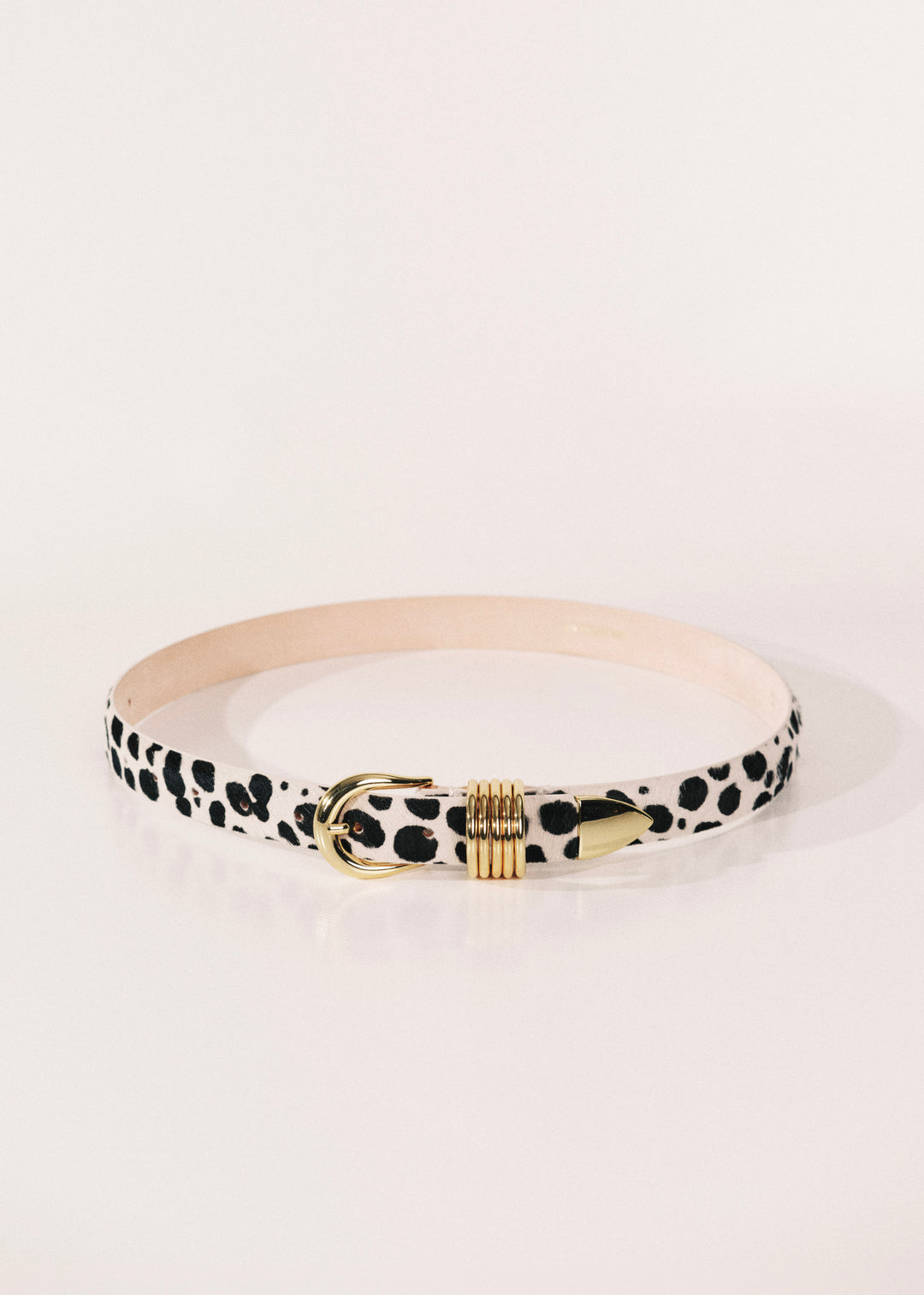 A slim belt with a black and white dalmatian print, featuring a gold-tone metal buckle and a beige interior lining. 