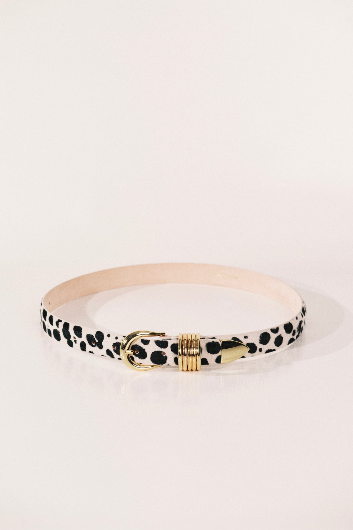A slim belt with a black and white dalmatian print, featuring a gold-tone metal buckle and a beige interior lining. 