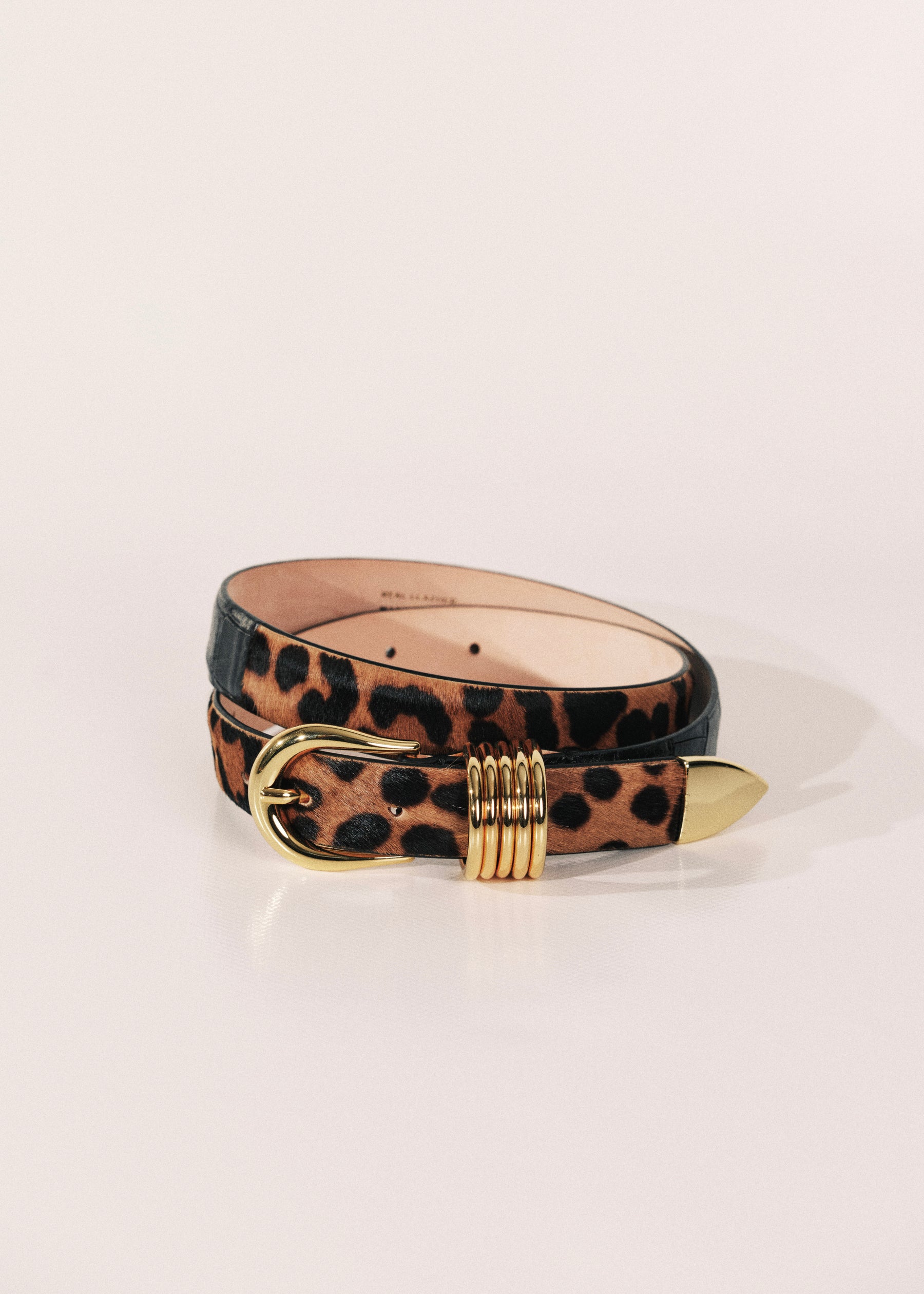 A belt featuring a leopard print pattern on the outer side and a smooth black leather inner side, finished with a gold-tone metal buckle and a decorative gold-tone ring detail on the tip.