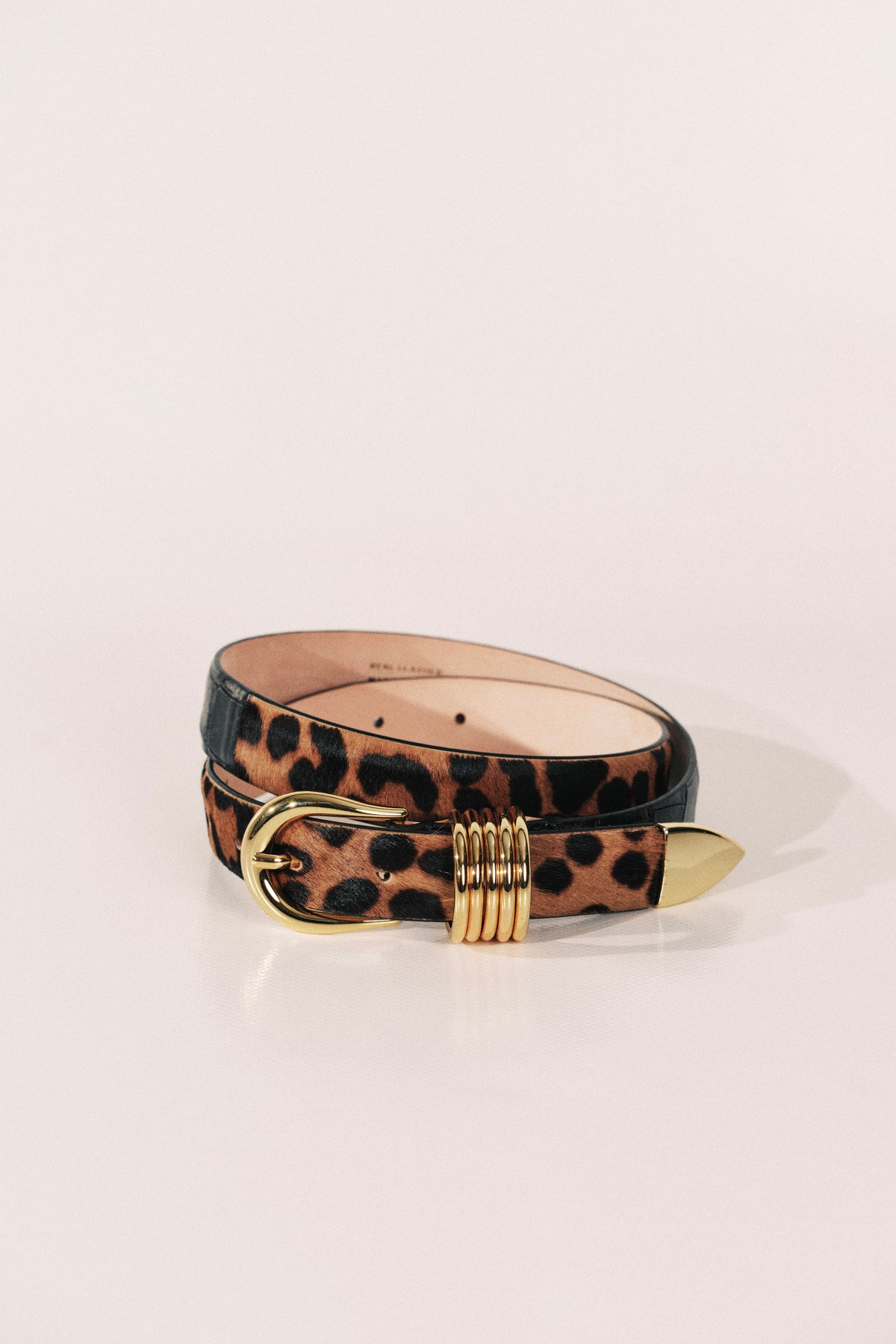 A belt featuring a leopard print pattern on the outer side and a smooth black leather inner side, finished with a gold-tone metal buckle and a decorative gold-tone ring detail on the tip.
