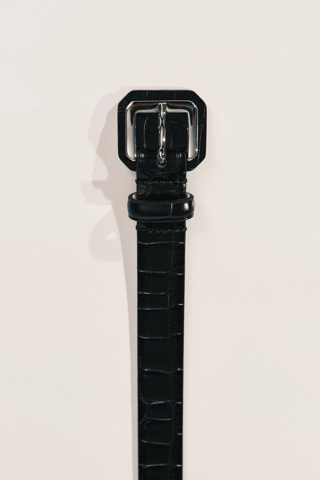A close-up of a black alligator-embossed leather belt with a silver metal buckle. 