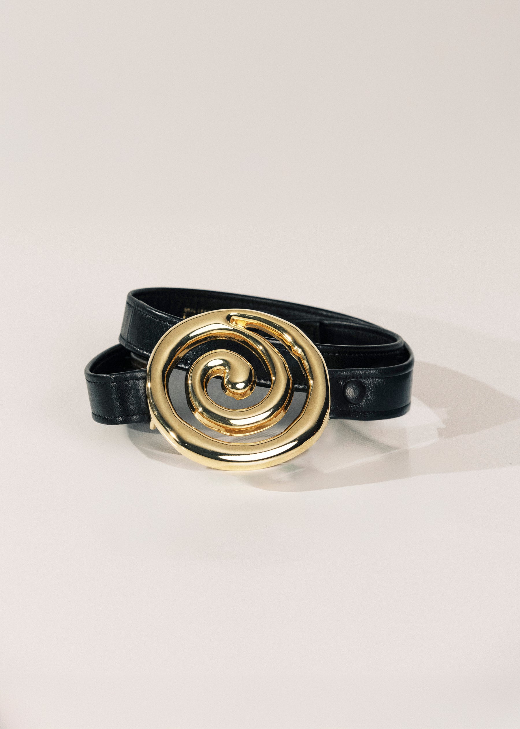 A black leather belt with a large, polished gold spiral buckle, displayed on a clean off-white background.