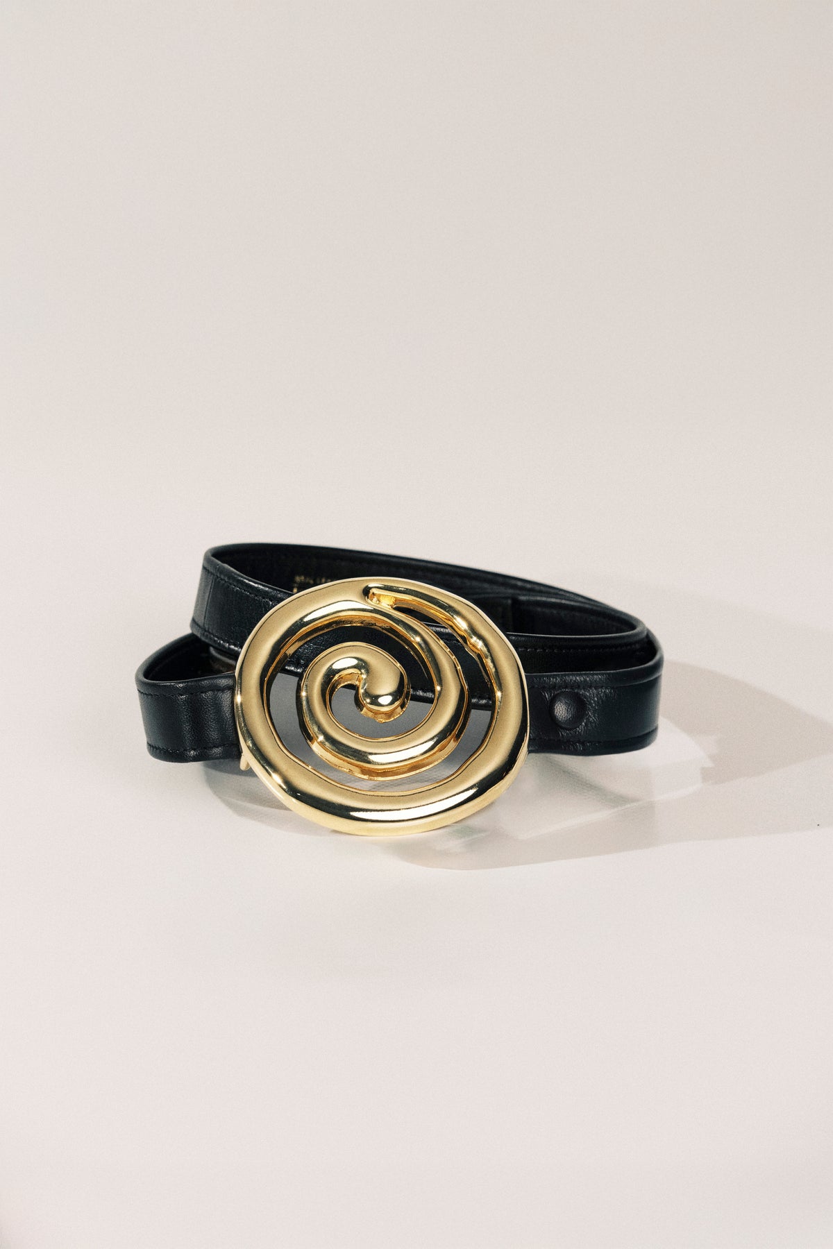 A black leather belt with a large, polished gold spiral buckle, displayed on a clean off-white background.