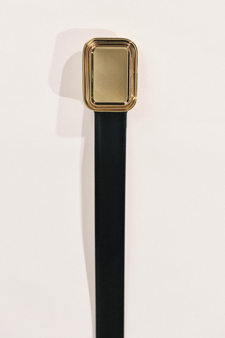 A close-up of a belt buckle with a rectangular gold-toned frame and a black strap, shown against a plain white background.
