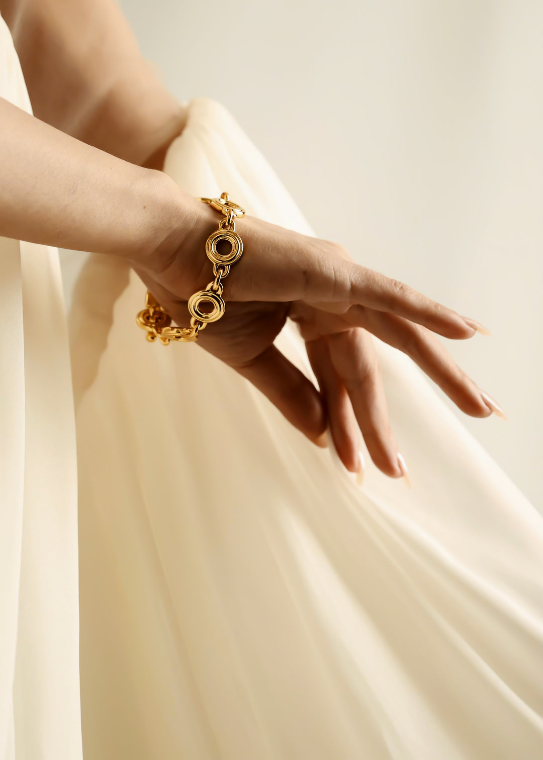 A close-up of a person's hand and wrist wearing a gold chain bracelet with circular links, posed against a soft cream-colored flowing fabric backdrop.