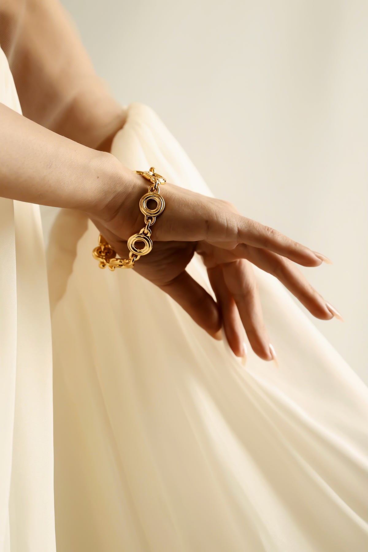 A close-up of a person's hand and wrist wearing a gold chain bracelet with circular links, posed against a soft cream-colored flowing fabric backdrop.