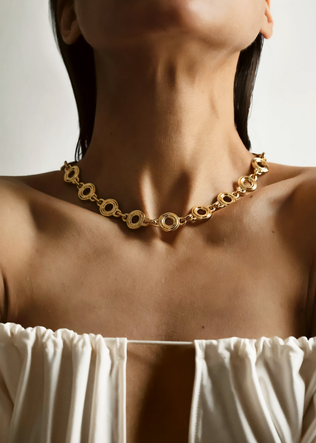 A close-up of a model wearing a chunky gold chain necklace with interlocking circular links, styled with a white off-the-shoulder top against a neutral background.