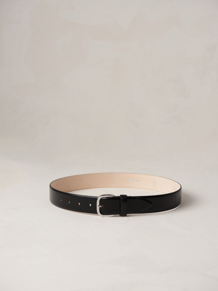 Déhanche jeanne belt in black leather with a silver buckle, elegantly coiled on a neutral background, showcasing high-quality craftsmanship and style.