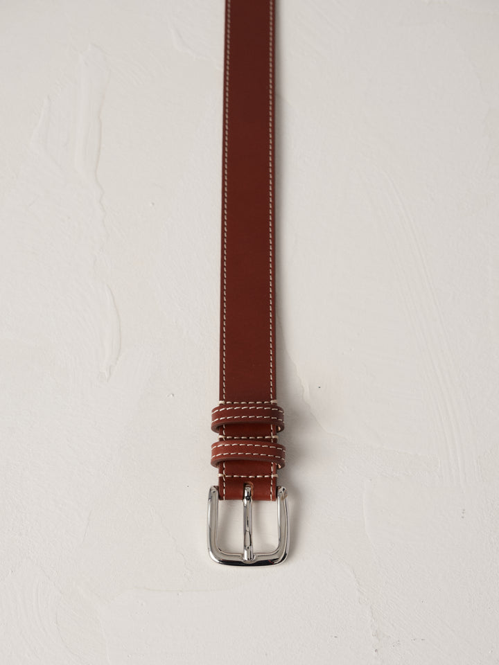 Déhanche louison belt in rich brown leather with white stitching and a silver buckle, elegantly coiled on a neutral background, showcasing refined craftsmanship.