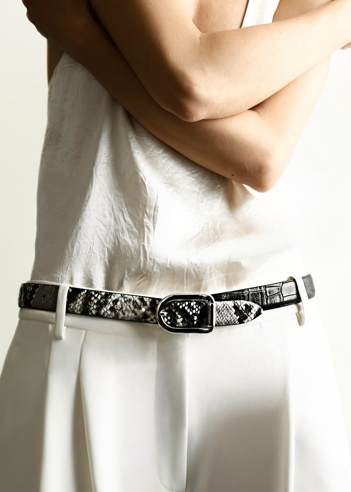 A model wearing a black and white snakeskin-patterned belt with a silver-tone buckle, paired with a white sleeveless top and white trousers.