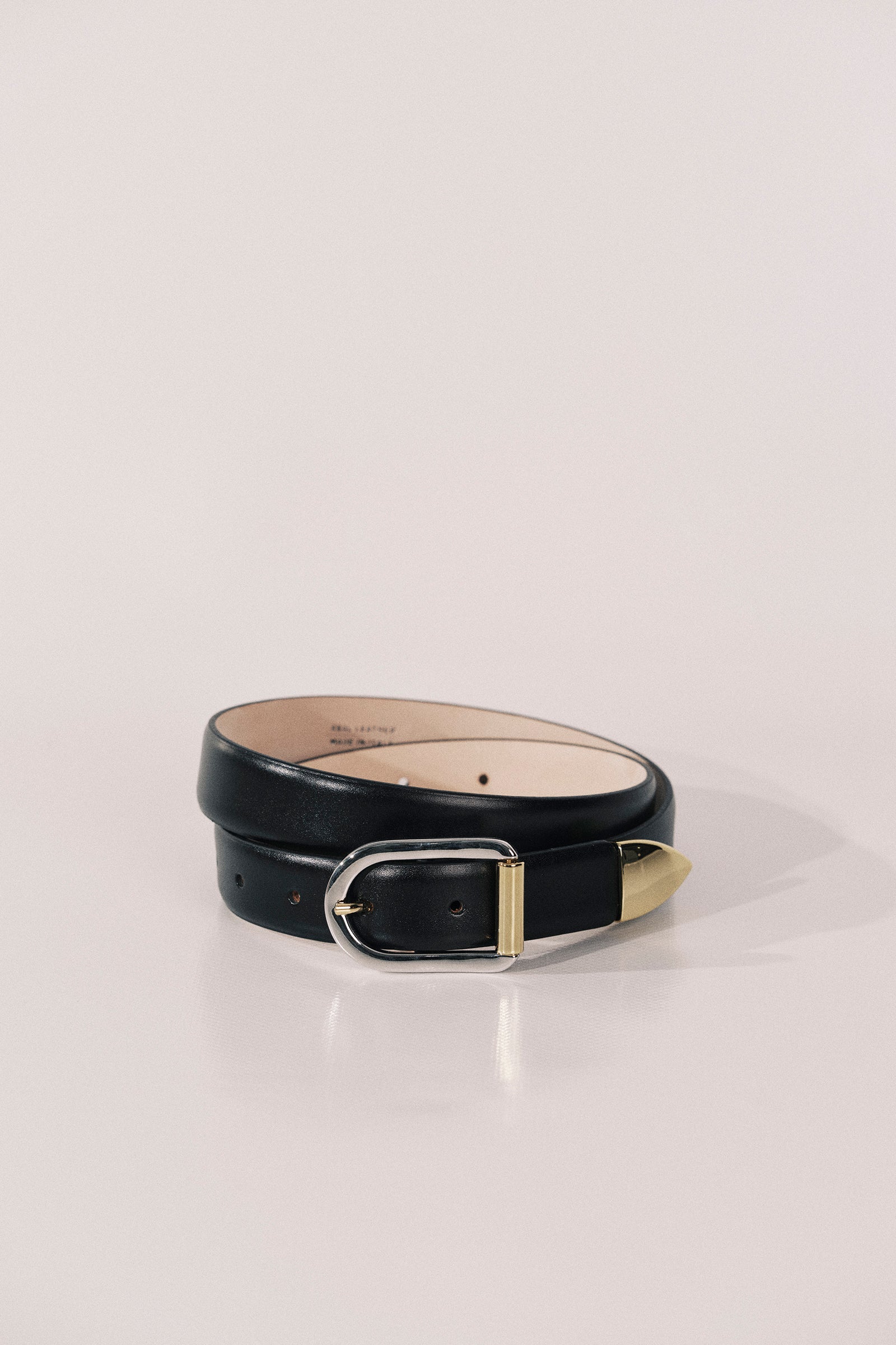 A black leather belt with a gold-tone metal buckle and tip, coiled on a light beige surface against a neutral background.
