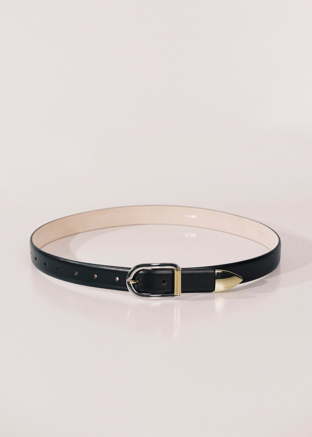 A slim black leather belt with a silver-tone buckle and gold-tone tip, displayed in a circular shape on a light cream background.