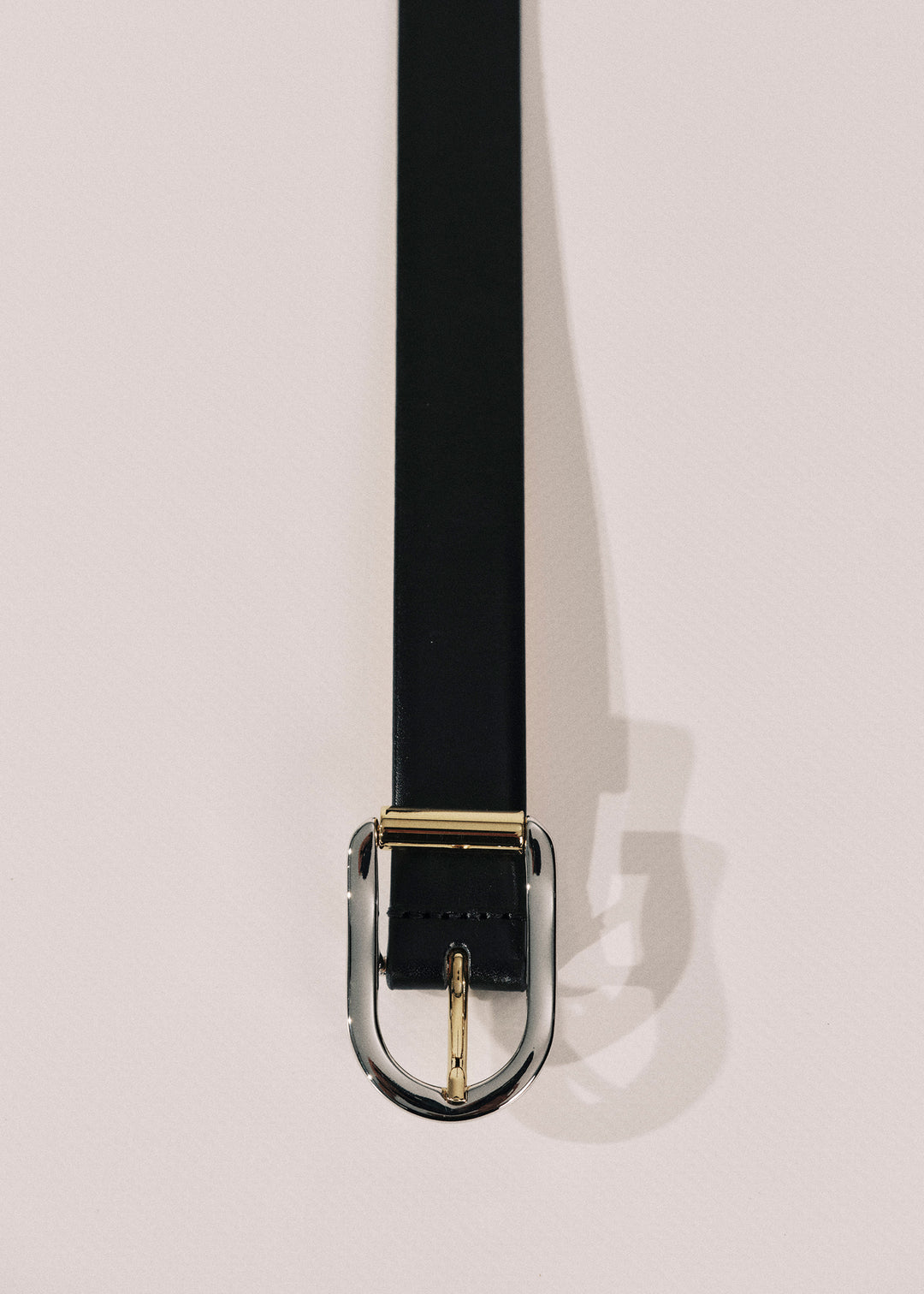 A black leather belt with a polished gold-tone metal buckle, shown hanging vertically against a light beige background.