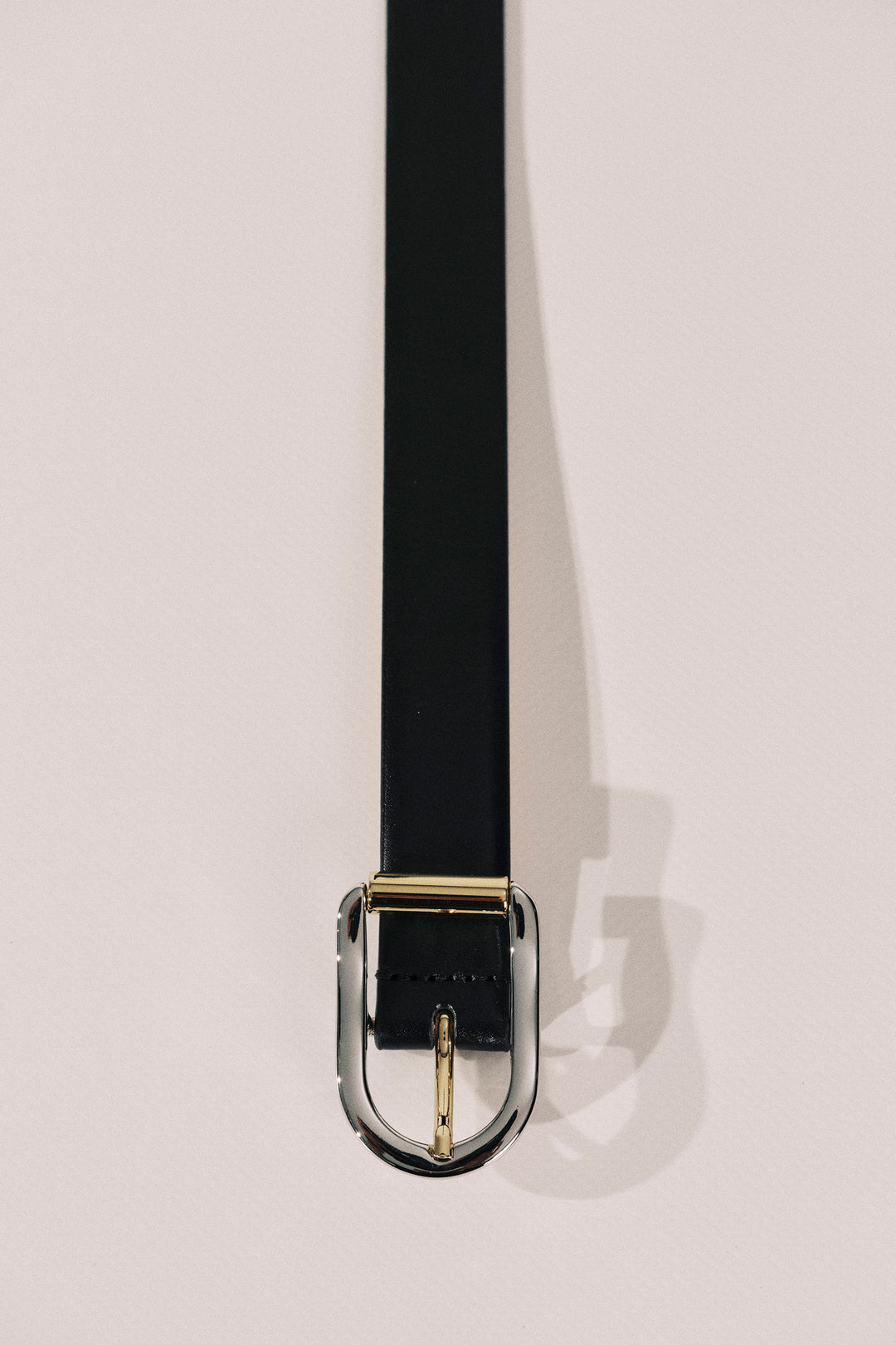 A black leather belt with a polished gold-tone metal buckle, shown hanging vertically against a light beige background.