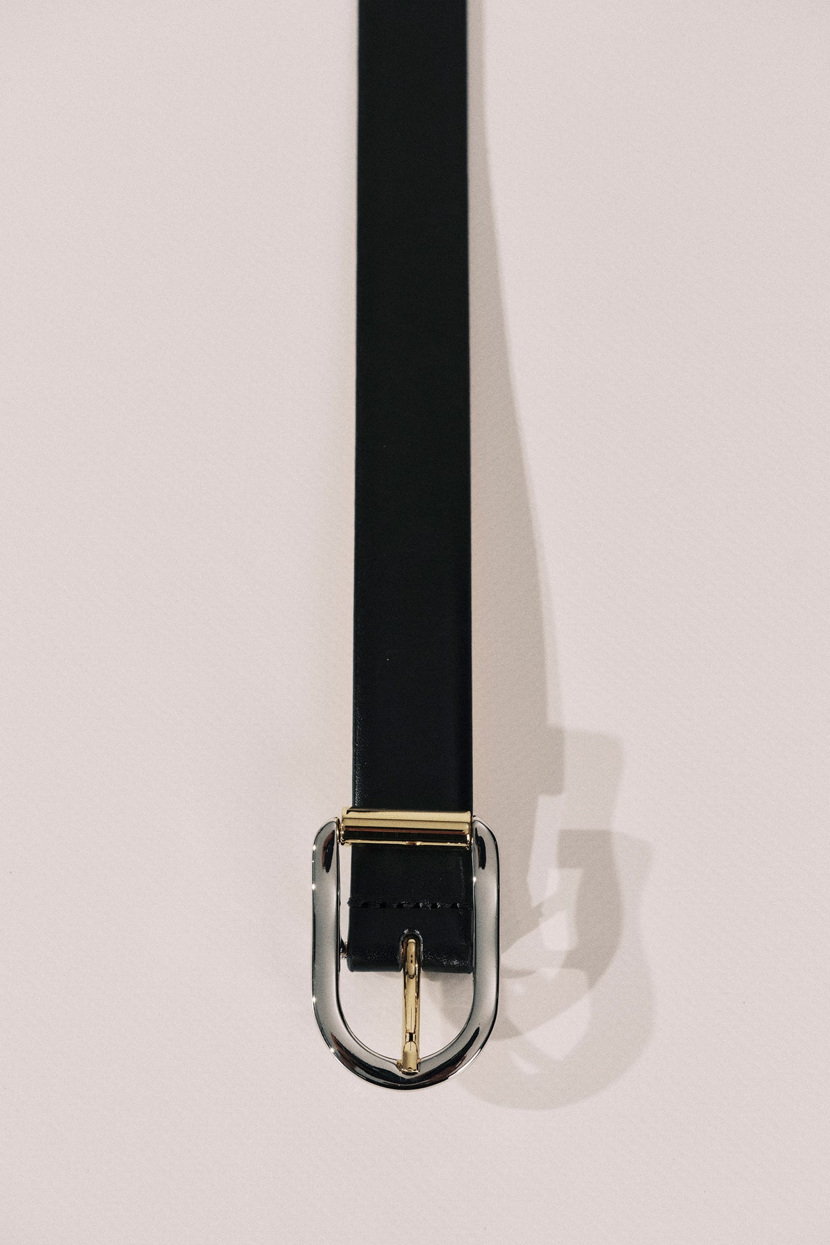 A black leather belt with a polished gold-tone metal buckle, shown hanging vertically against a light beige background.