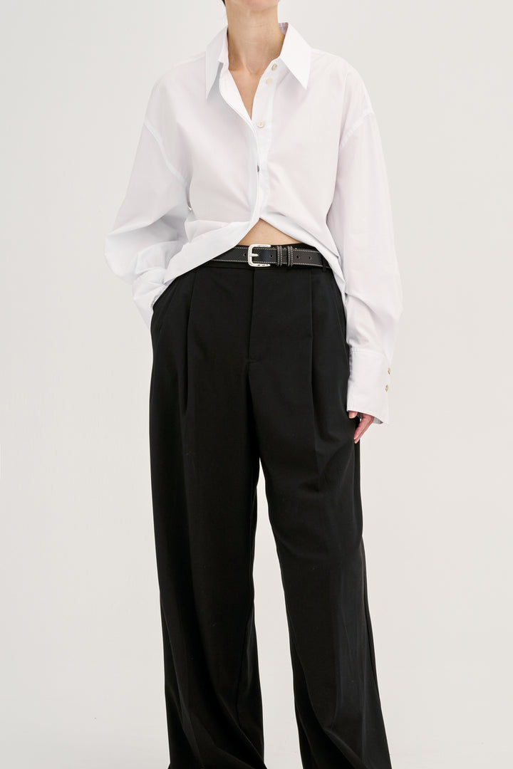 Model wearing Déhanche Louison black leather belt with white stitching and silver buckle, paired with black trousers and a white shirt.