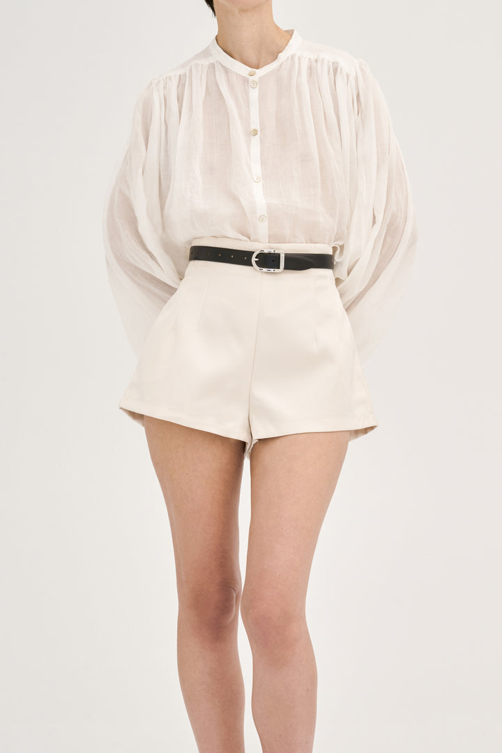 Model wearing Déhanche Mija black leather belt with silver buckle, paired with high-waisted beige shorts and a sheer white blouse.