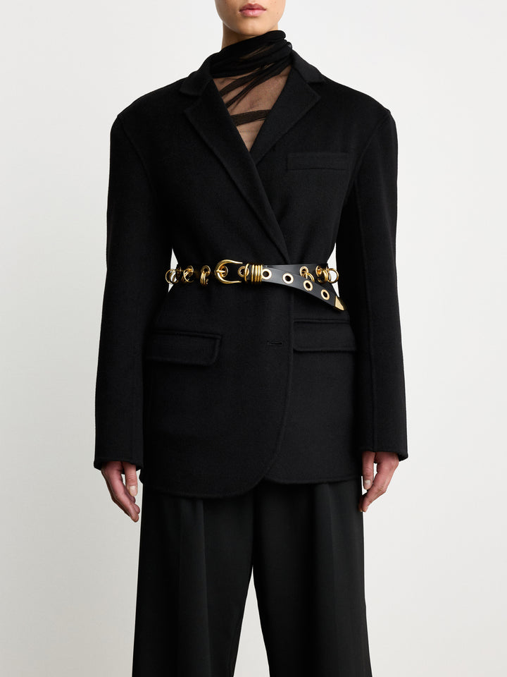 Model wearing Déhanche Revenge Gold black leather belt with gold grommets and buckle, styled over a black blazer and black trousers.