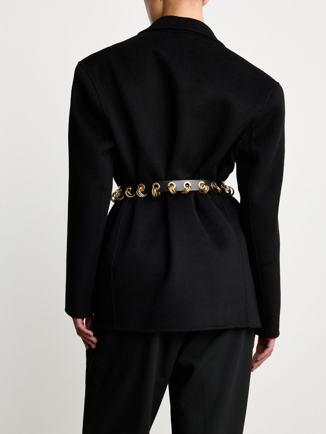 Model wearing Déhanche Revenge Gold black leather belt with gold grommets and buckle, styled with a black blazer and black trousers, back view.