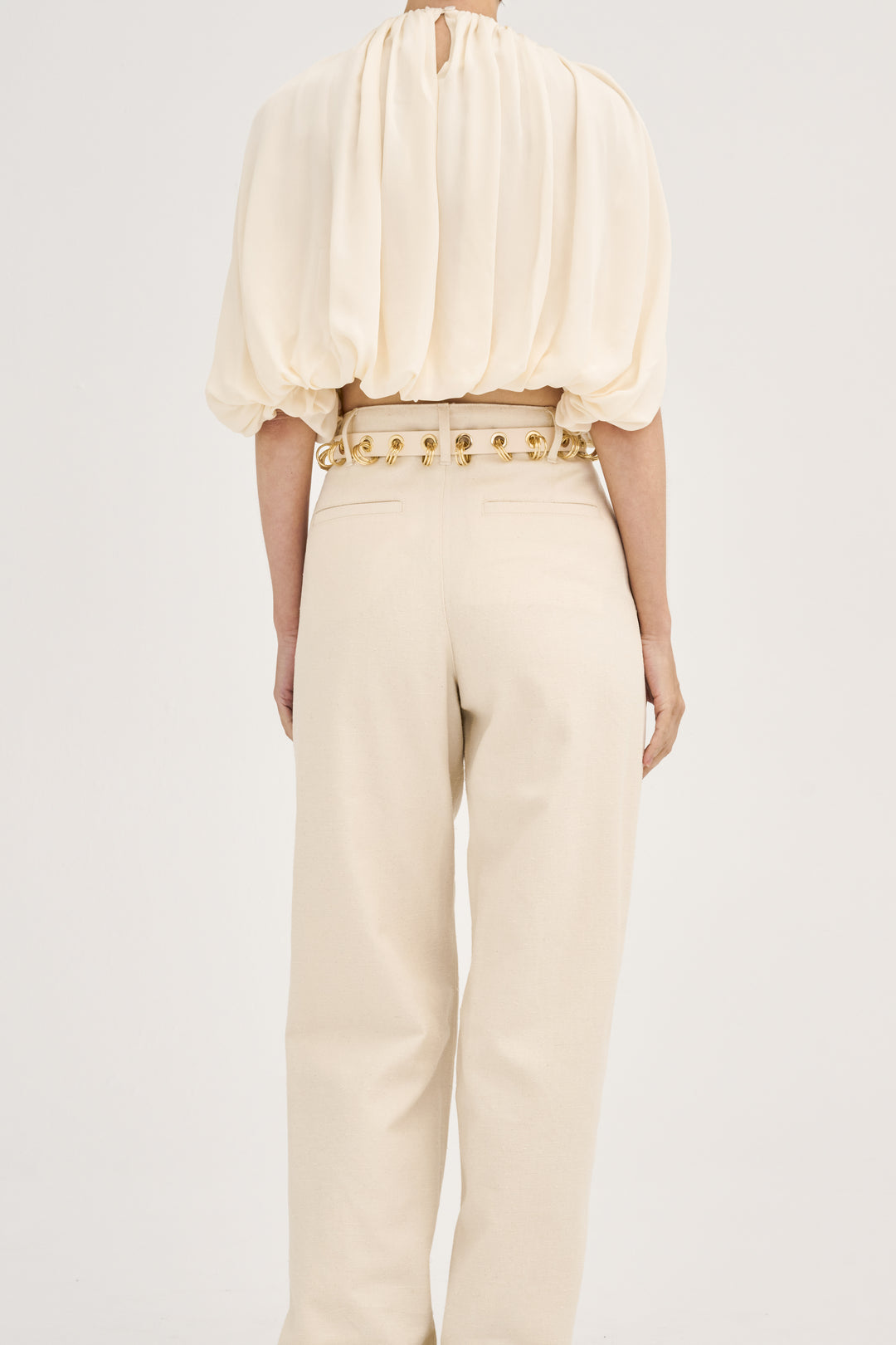 Model wearing Déhanche Revenge Gold white leather belt with gold grommets and buckle, styled with high-waisted cream pants and a flowy cream blouse, back view.