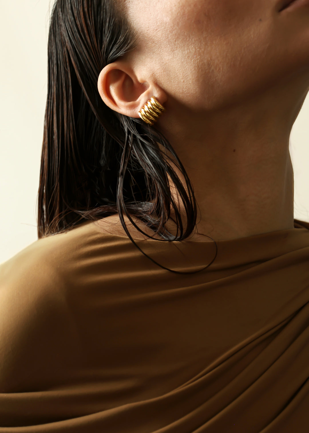 A close-up of a model wearing a gold earring, with wet dark hair and a draped brown top against a light beige background.