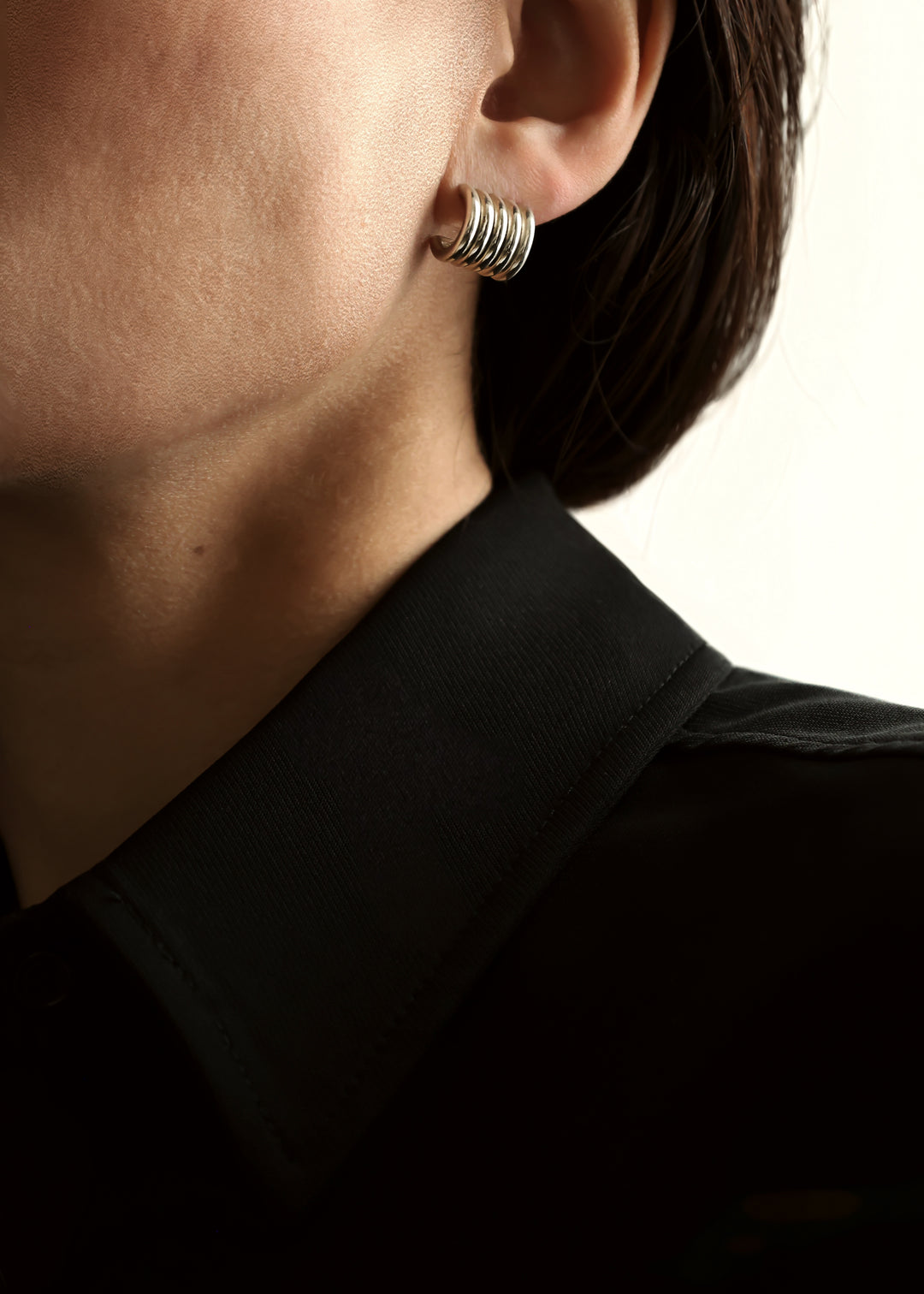 A close-up of a person's ear wearing a small silver earring with multiple concentric rings, paired with a black collared shirt against a light background.