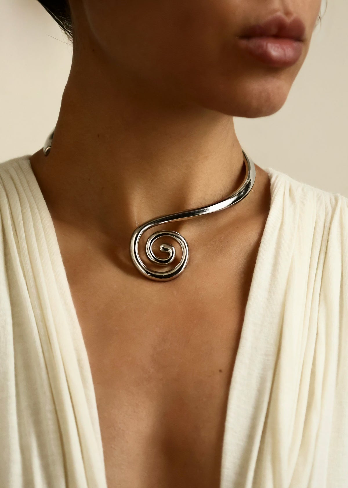 A close-up of a model wearing a polished silver spiral choker necklace with a wide, draped cream-colored top.