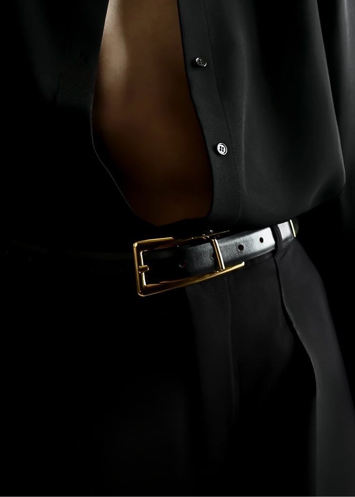 A close-up of a black leather belt with a gold-tone rectangular buckle worn at the waist over a black shirt and trousers.