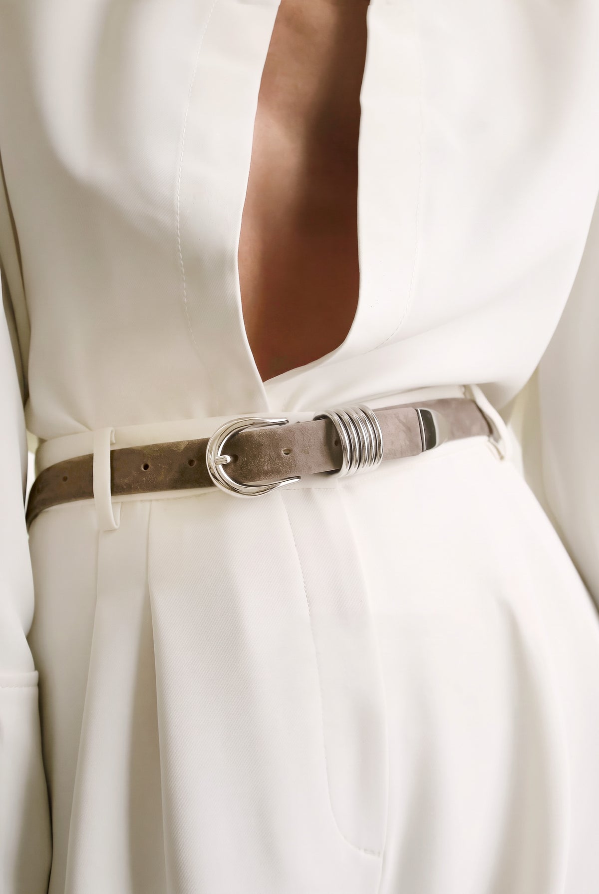 A close-up of a person wearing a white shirt and pants, styled with a taupe-grey suede belt featuring silver hardware