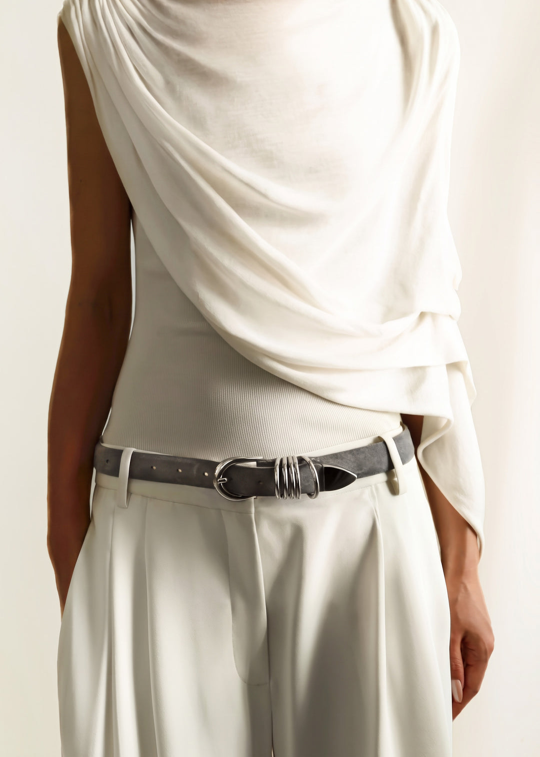 A close-up of a model wearing a draped off-white top with matching draped trousers and a fog grey suede belt with silver hardware.