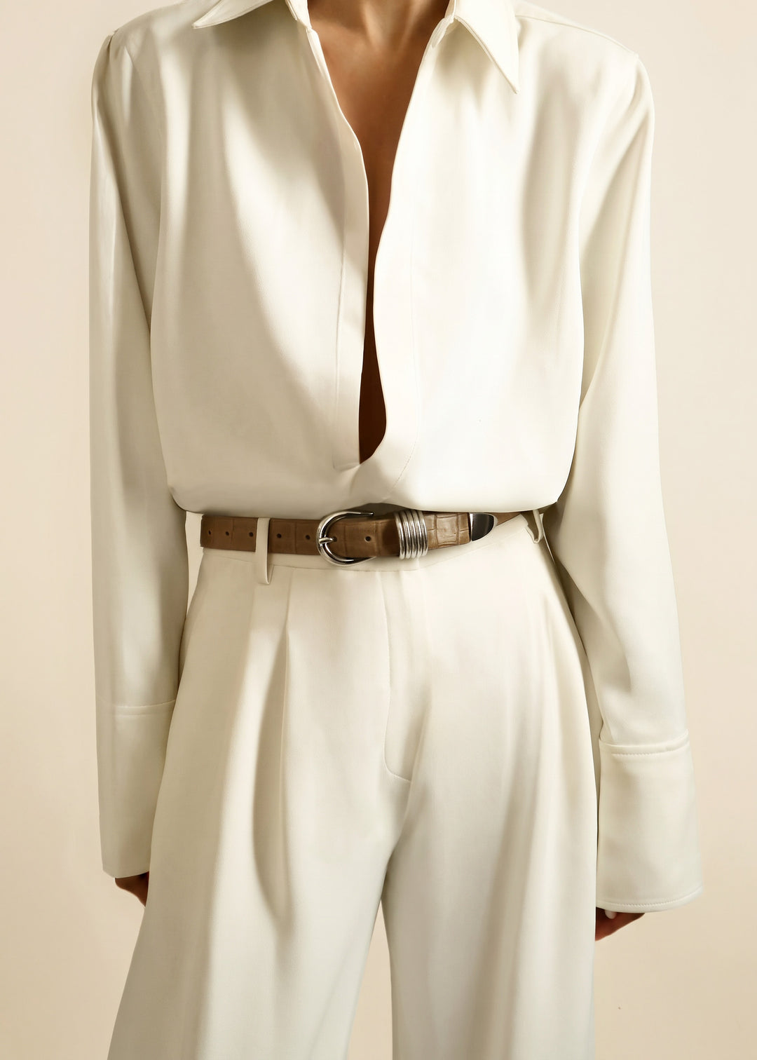 A model wearing a cream-colored long-sleeve button-down shirt tucked into matching cream trousers, styled with a chai brown alligator leather belt featuring silver hardware.