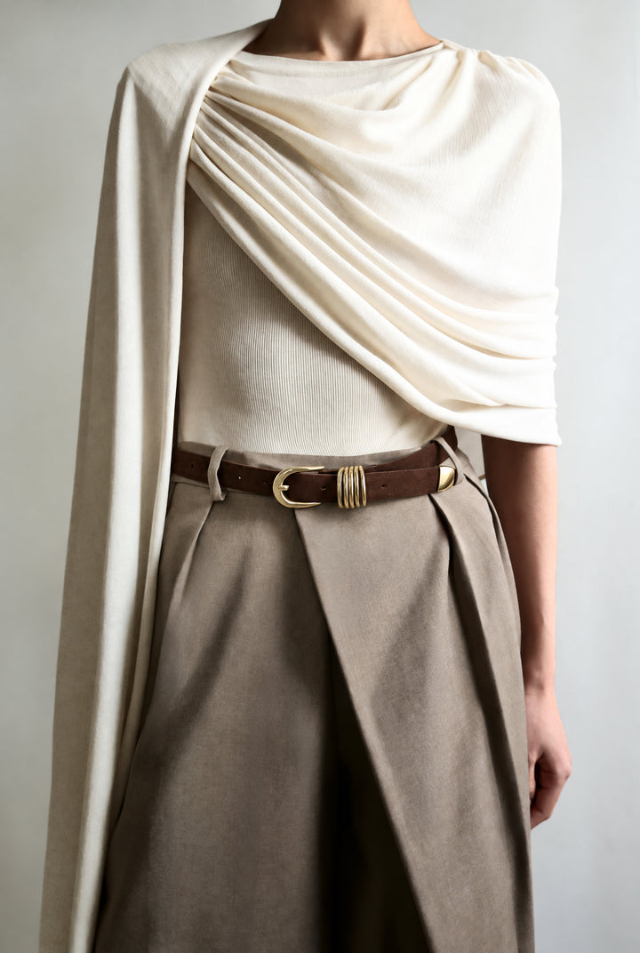 A model wearing a cream draped knit top with a matching draped capelet, paired with pleated taupe trousers and a brown suede belt with gold hardware.