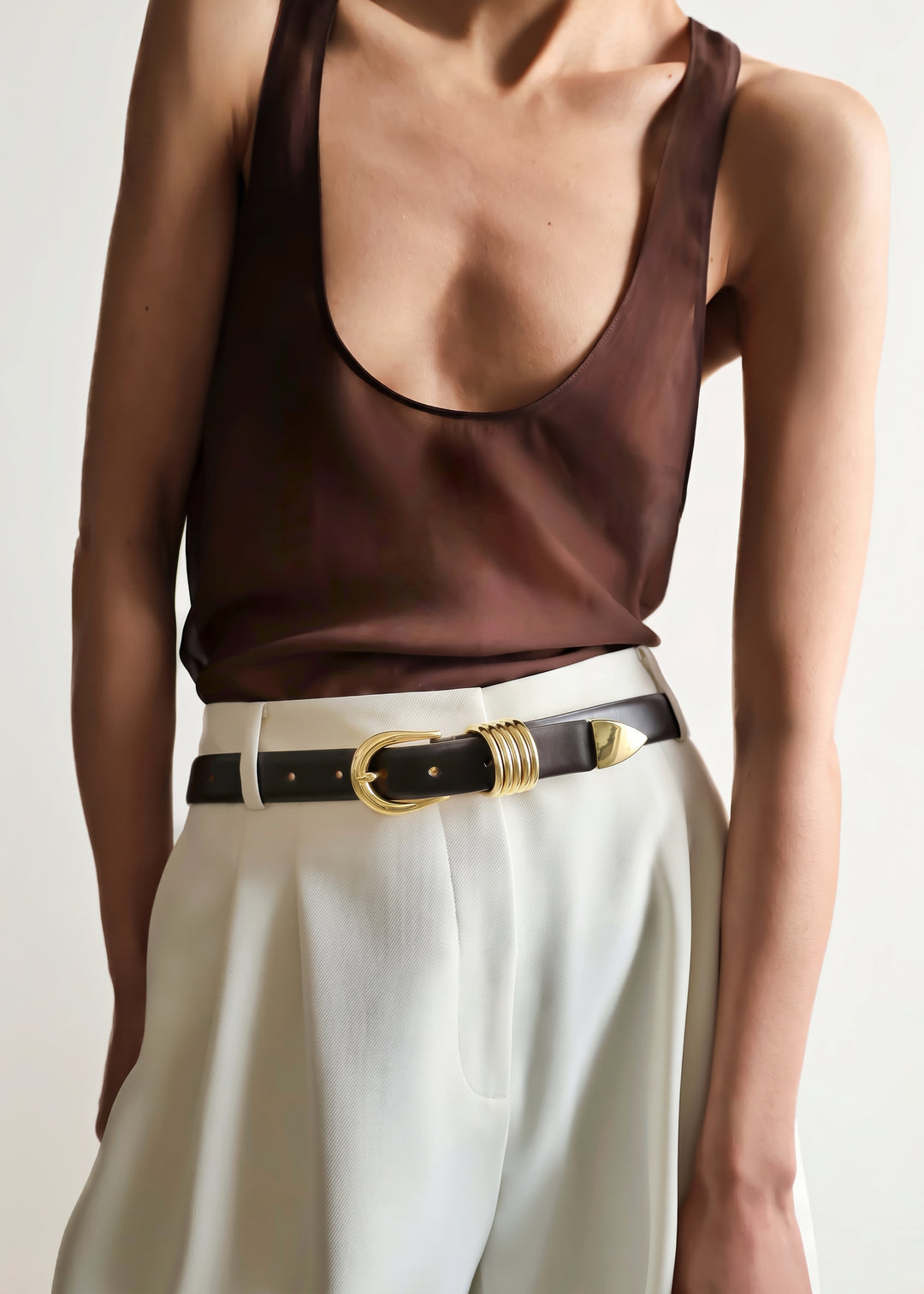 A close-up of a model wearing a dark brown satin tank top and white trousers, styled with a purple marrone leather belt featuring gold hardware