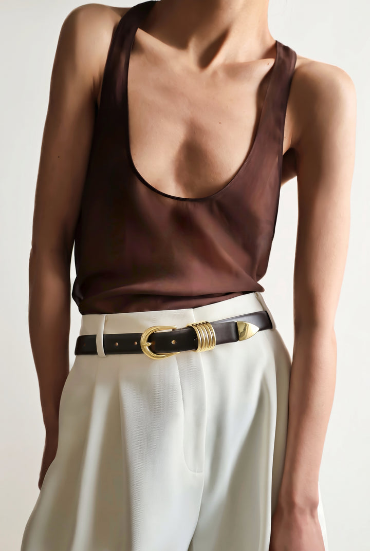 A close-up of a model wearing a dark brown satin tank top and white trousers, styled with a purple marrone leather belt featuring gold hardware
