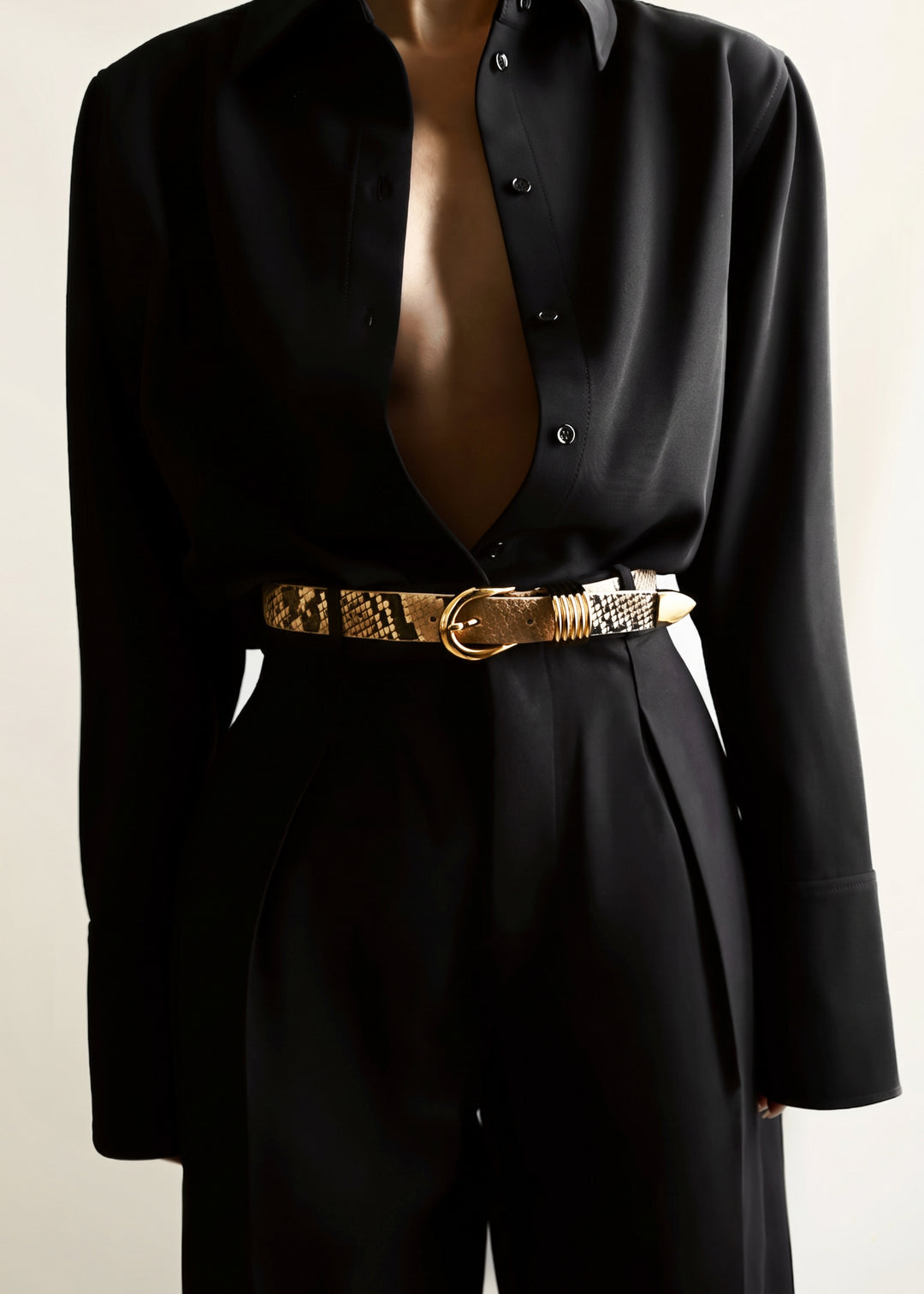 A close-up of a model wearing a black long-sleeve button-up shirt and matching black trousers, styled with a textured gold and black snakeskin-patterned belt with a gold buckle.