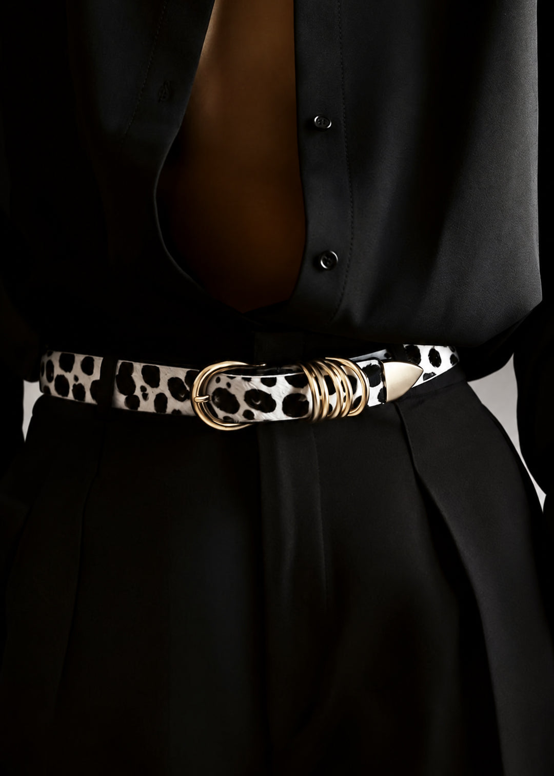 A close-up of a model wearing a black button-down shirt and black pleated skirt, accessorized with a black and white dalmatian print belt with gold hardware