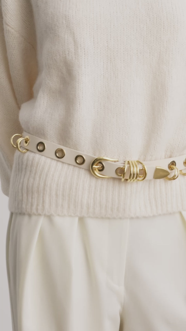 Model wearing Déhanche Revenge Gold white leather belt with gold grommets and buckle, styled with cream trousers and a cream sweater.