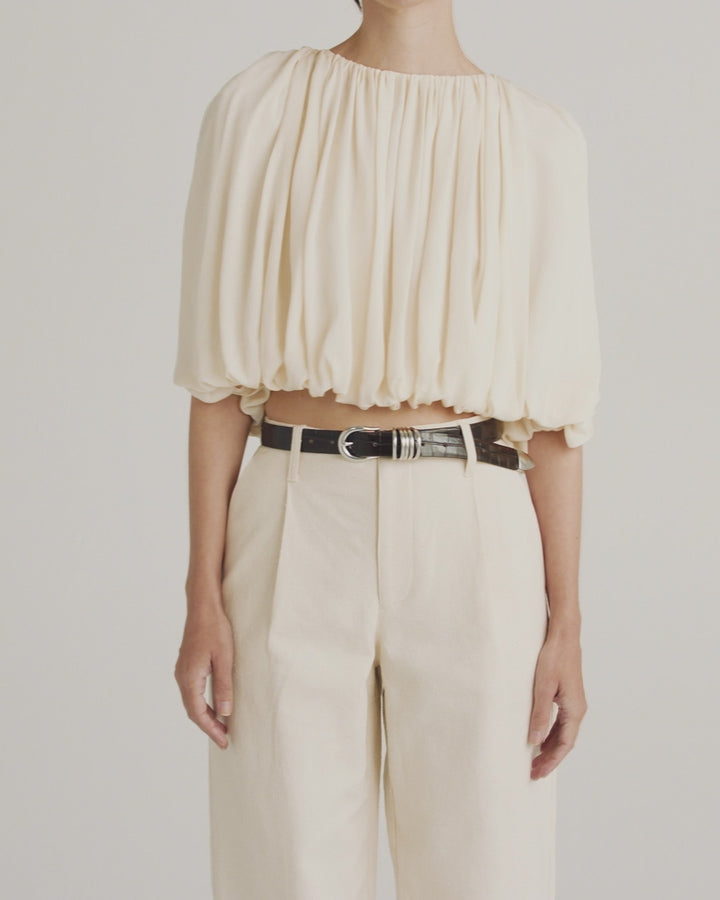 Déhanche Hollyhock Croco belt in black crocodile leather, paired with a voluminous ivory blouse and cream trousers, showcasing sophisticated style.