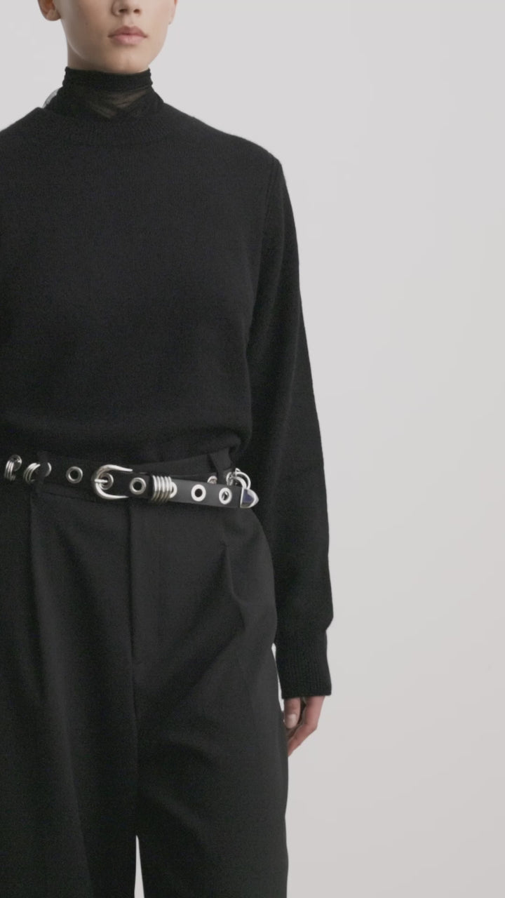 Déhanche Revenge Silver belt in black leather with silver grommets and buckle, worn with black high-waisted pants and a black sweater.