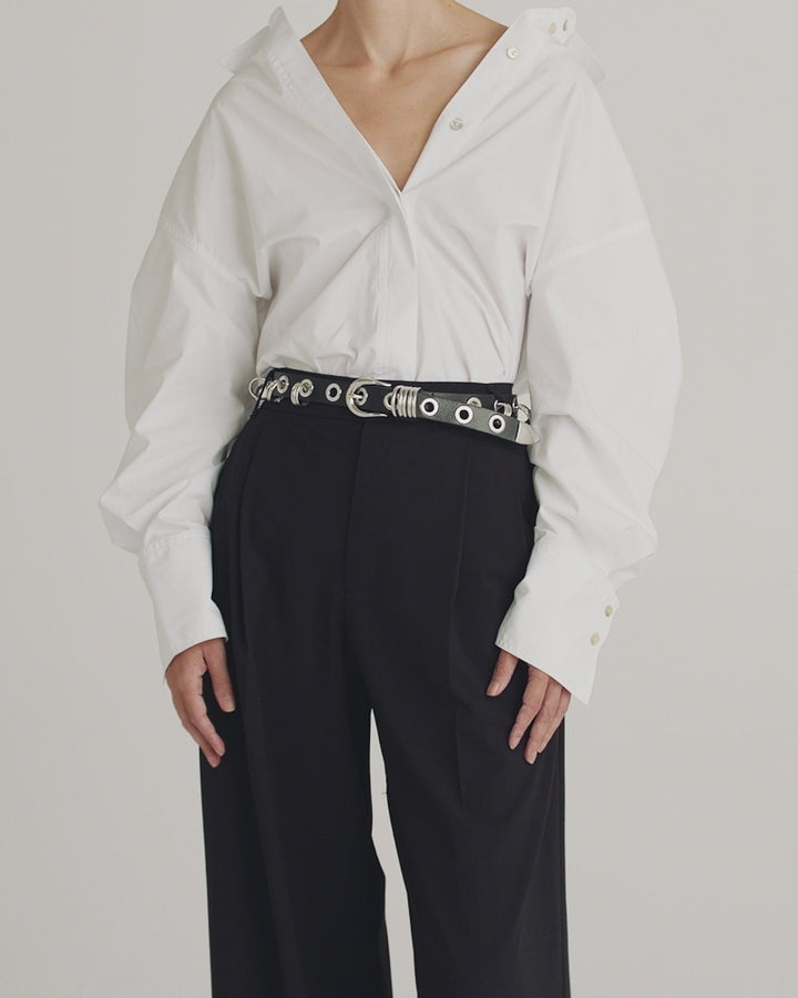 Déhanche Revenge Silver belt in black leather with silver grommets and buckle, paired with a white oversized shirt and black trousers.