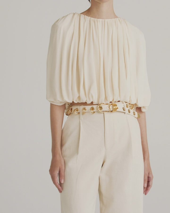 Model wearing Déhanche Revenge Gold white leather belt with gold grommets and buckle, styled over a cream blouse and matching pants.
