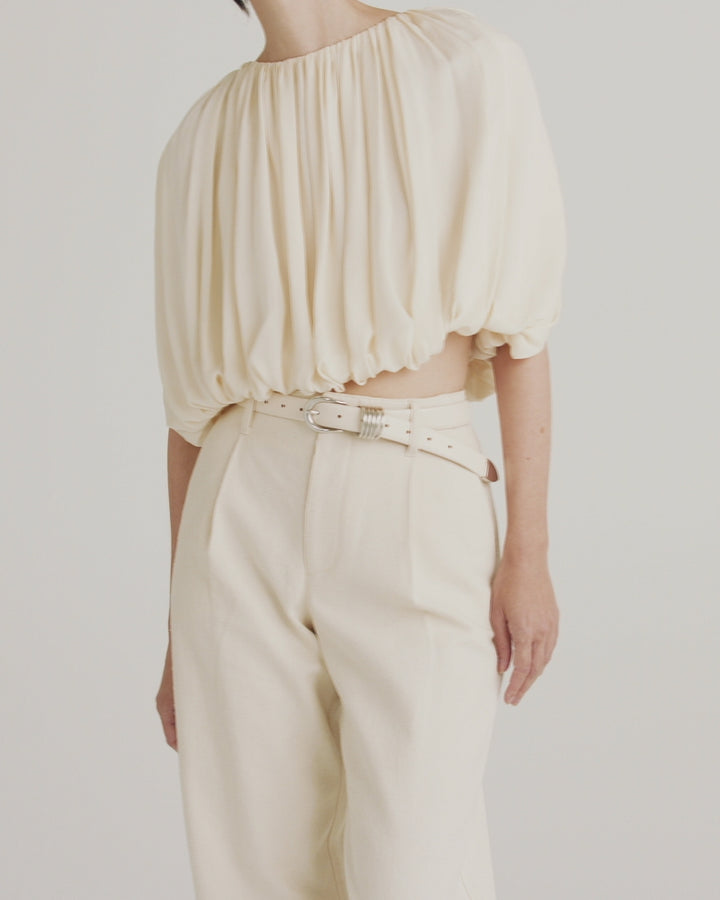 Model showcasing Déhanche Hollyhock belt in cream leather with a silver buckle, paired with an elegant, gathered cream blouse and matching high-waisted trousers for a refined, monochromatic look.