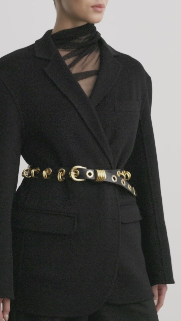 Model wearing Déhanche Revenge Gold black leather belt with gold grommets and buckle, styled over a black blazer and black trousers.