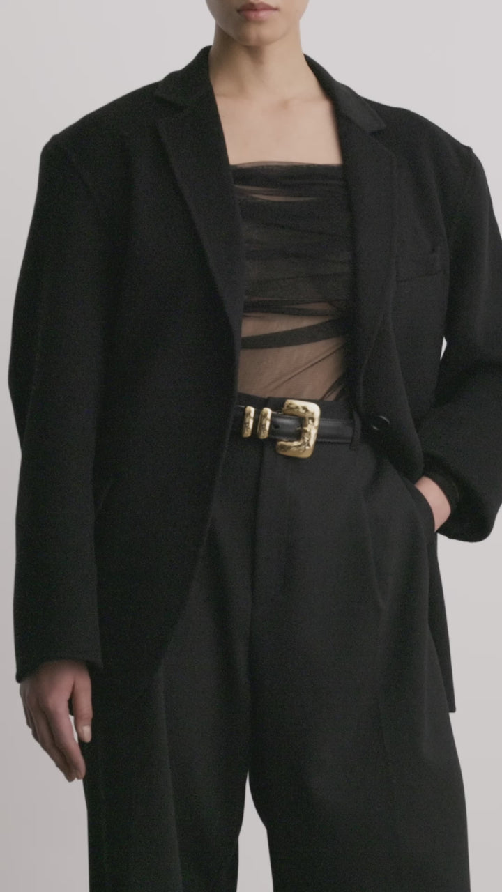 Déhanche Tetra belt in black leather with a distinctive gold buckle, worn with a black halter top and high-waisted black pantsDéhanche Tetra belt in black leather with a distinctive gold buckle, paired with a sheer black off-the-shoulder top and high-waisted black pants.