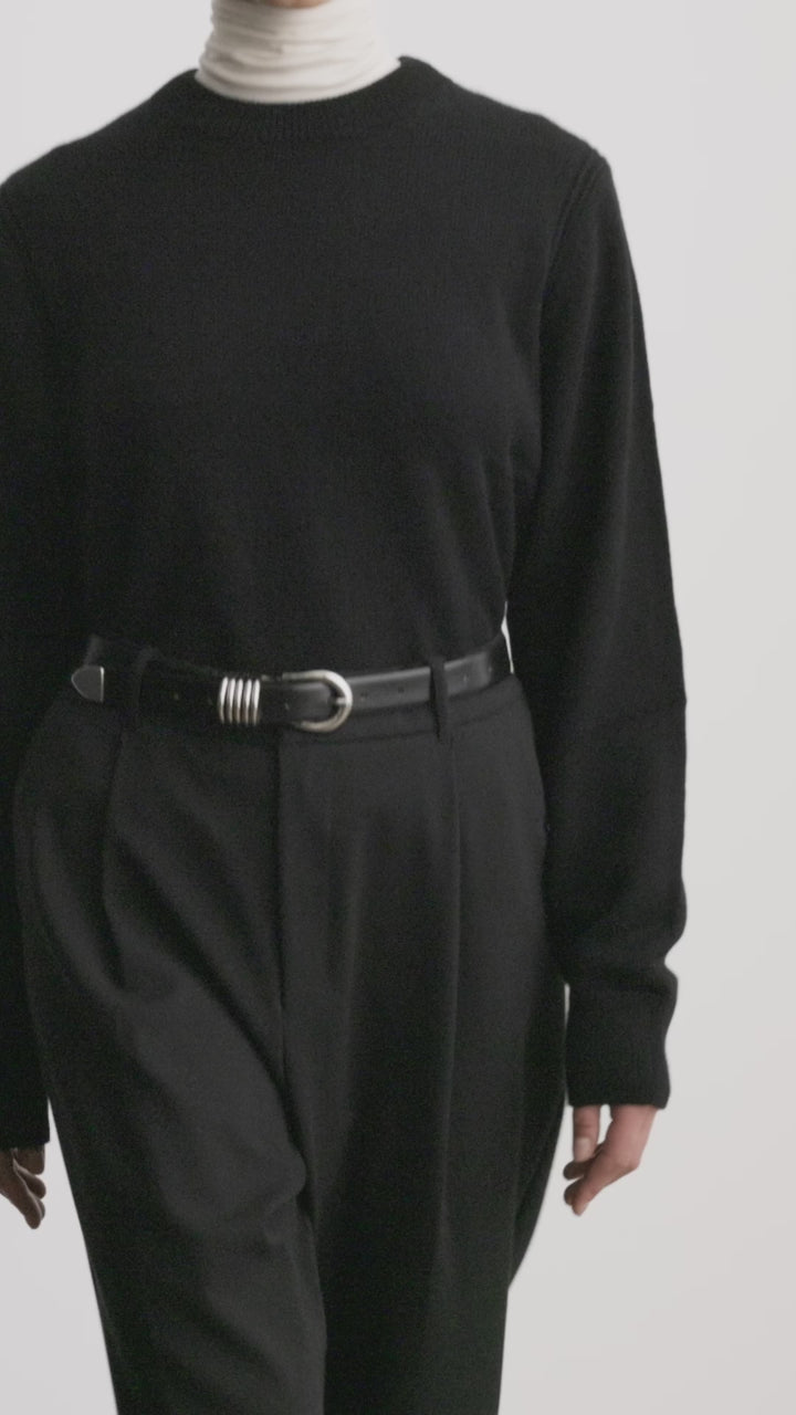 Model wearing Déhanche Hollyhock belt in sleek black leather with an intricate silver buckle, paired with a black sweater and high-waisted trousers, showcasing modern elegance and sophistication.